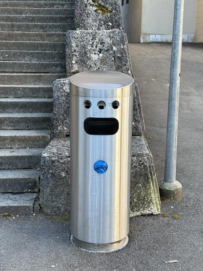 Ein Mülleimer in der Stadt Solothurn. Er besteht aus einer runden, blanken Stahltonne mit einer ovalen, länglichen, und quer liegenden Öffnung für Müll. Darüber befinden sich drei kleine, kreisrunde Öffnungen für Zigaretten. Weiter unten ist eine weitere kreisrunde Öffnung, der man Plastiksäckchen für Hundekot entnehmen kann.