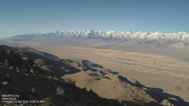 A view of the Owens Valley looking south. In the foreground, rocks and vegetation can be seen on the slopes. The valley below is a variety of tans and browns. Rising in the distance are snow-covered mountains. The sky above is blue and clear.