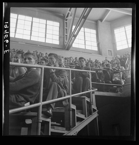The image is a black and white photograph showing an audience in what appears to be an indoor arena or auditorium. Several young men are seated on tiered metal bleachers, attentively listening as they watch something off-camera that has captured their attention. The windows behind them let natural light into the space, highlighting the rows of people below.

The photo is marked with handwritten notations "7-13" and possibly a location or date reference in another language at its top left corner. It captures a moment of collective focus among what appears to be university students during an event involving General Smedley Butler on Peace Day, as indicated by the caption from Student audience listening to Peace Day address of General Smedley Butler.

The overall atmosphere is one of engagement and contemplation, with individuals absorbed in their surroundings or possibly reflecting upon a speech they have heard.