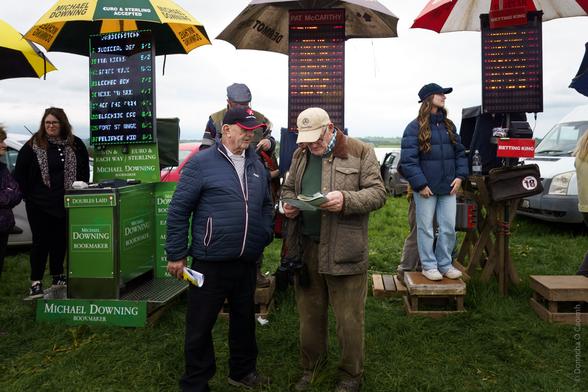 A lively scene at a rural Irish racecourse featuring bookmakers' stands under colourful umbrellas, including visible signage such as "Michael Downing Bookmaker," "Pat McCarthy," and "Betting King." Two men are engaged in conversation, one holding a betting slip, while others stand nearby on wooden platforms. The setting is grassy with overcast skies.