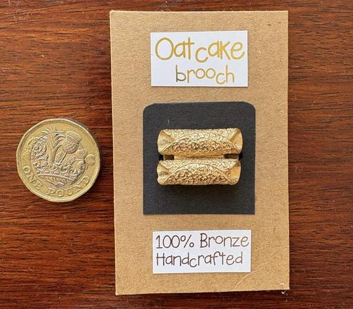 Photo of a small goldish brooch in the shape of two rolled up oatcakes side by side. It is attached to a black square on a larger brown card with the legend "Oatcake brooch - 100% Bronze Handcrafted". A £1 coin sits alongside for scale. Both items are against a brown wooden background.