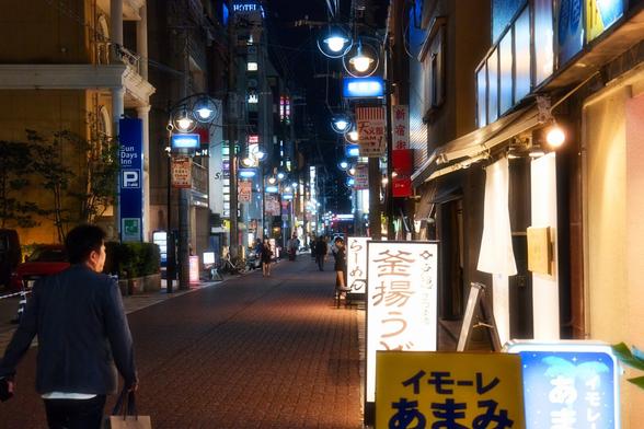 Taken close to the bright lights of a street restaurant on a busy throughfare in night-time Kagoshima.
Pools of light are cast onto the pavement and the whole street is flecked with lights and street signs.