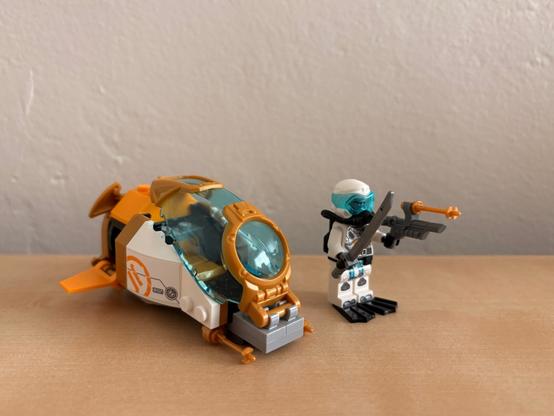 LEGO space-themed set featuring a small orange and white spacecraft or pod with a transparent blue cockpit alongside a minifigure astronaut. The astronaut wears a white helmet with blue visor, white space suit with black and blue detailing, and carries what appears to be a weapon or tool. Both items are displayed on a light brown surface against a neutral beige wall background.​​​​​​​​​​​​​​​​