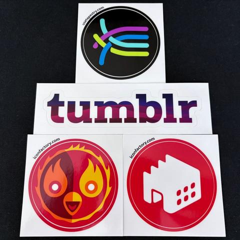 Four stickers against a black background. The top sticker shows a colorful circular logo with curved lines in teal, purple, blue, and lime green on a black background. Below it is a white "tumblr" sticker with gradient text in purple and pink. The bottom two are circular red stickers: left shows a cartoon flame bird character with eyes and a smile, right displays a white app or media player icon. All stickers have "iconfactory.com" printed in small text.