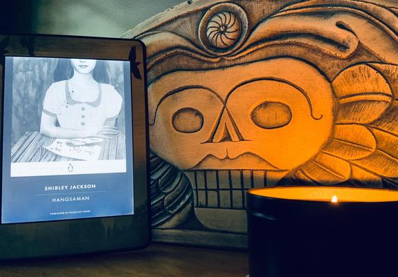 Pictured is a shadowed photo of a candle reflecting light onto a carving of a winged skull (inspired by an old New England headstone). In the corner is an ebook reader showing the cover of Shirley Jackson’s novel Hangsaman.
