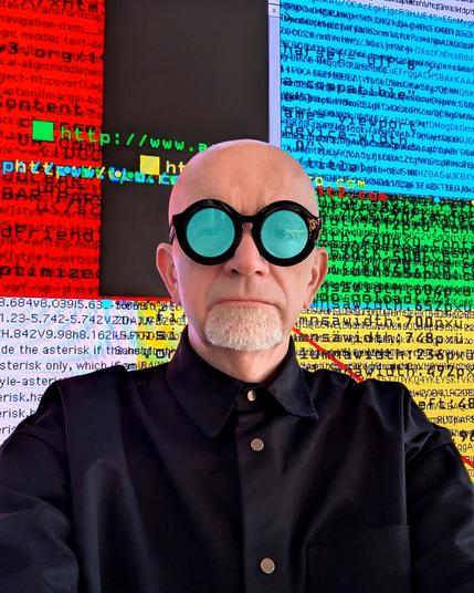 A bald man with a white goatee, wearing black round sunglasses with turquoise lenses and a high-collared black shirt with metallic buttons. Behind him, a large screen displays a chaotic mix of colorful, glitched text, code, and pixelated graphics in red, yellow, blue, green, and black. The digital distortion suggests themes of internet history, coding, or digital art.