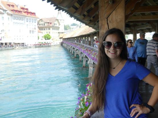 Girl with long brown hair and blue tee shirt posing with left hand on hip on chapel bridge on lake lucerne in luzern switzerland in sunglasses. Emily Grice.