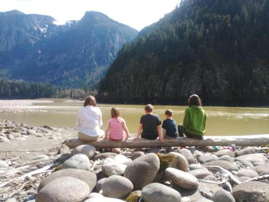 A group of five people, are sitting on a large log by the edge of a river, facing away from the camera. The three in the middle are younger children. The river is surrounded by rocky terrain and mountains covered in trees. The sky is partly cloudy.