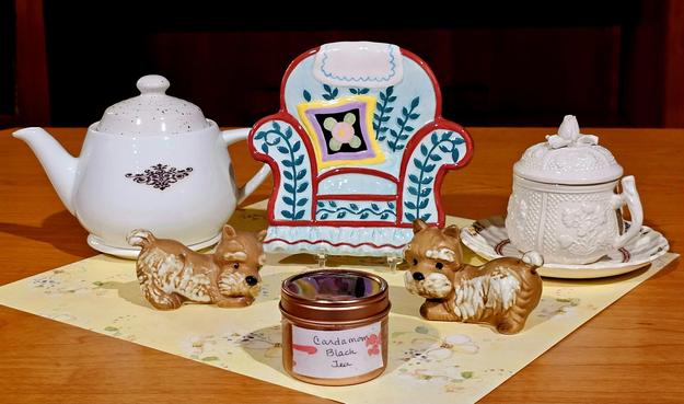 Tea tableau of two puppies playing in a parlor. On a flowered yellow mat are a teapot and a lidded cup and saucer with a colorful ceramic armchair in between them. In front of the chair are two porcelain tan terriers bowing playfully at each other. The tea is Cardamom black loose leaf tea from Sadif.