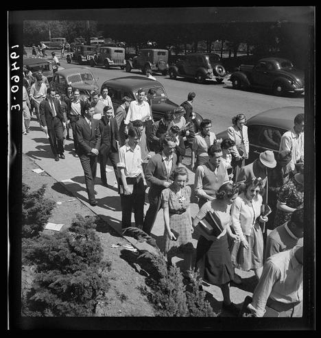 The image is a black and white photograph depicting a large group of people, predominantly young adults dressed in mid-20th-century attire, walking down what appears to be a city street or pedestrian path. They are moving towards the right side of the frame with some individuals looking back as if they're aware that their picture is being taken.

The crowd comprises men and women wearing various styles such as suits for men and dresses or skirts and blouses for women. Some people carry books, suggesting an academic context. The attire suggests a time period likely between the 1940s to early '50s based on fashion trends of that era.

On one side of the path are parked vehicles including cars and trucks typical of mid-century design, indicating this event is taking place in an urban area with sufficient road space for vehicular traffic. On the opposite side from where people walk appears a row of trees or shrubs which separate them from what could be residential properties or another public pathway.

The photograph captures a sense of organized movement and unity among the crowd, possibly indicative of participation in a specific event like a march or parade. The text '3:02pm' visible at the top left suggests that this was captured during daylight hours on March 1st, but no year is provided within the image.

The overall scene conveys an atmosphere of communal activity and shared purpose among individuals who seem to be en [...]