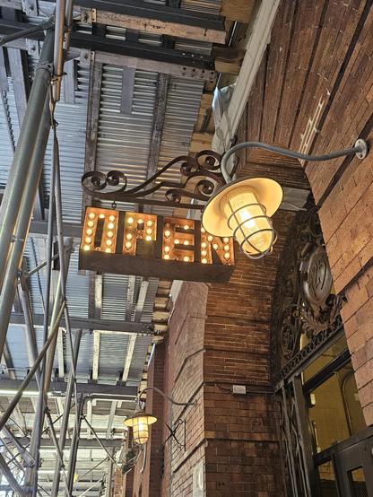Photograph os a sign saying 'OPEN' with incandescent bulbz hung from s wrought iron curly decorative arm, infront of a cage,protected work light. On a thin redbrick building
