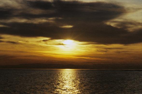 A photo of a sunset over the sea, dark clouds are partially obscuring the setting sun, everything has an orange tint to it