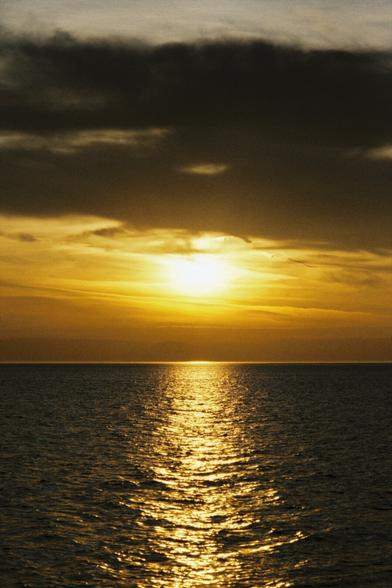 A portrait image of a sunset over the sea, dark clouds are partially obscuring the setting sun, everything has an orange tint to it
