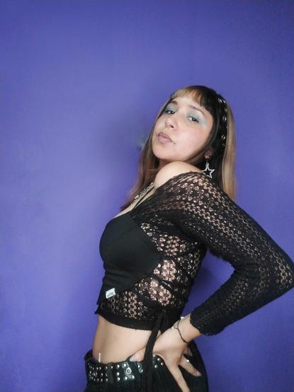Una joven con cabello rubio y negro con flequillo, luce un maquillaje con sombras de ojos azules. Lleva una diadema negra con remaches y un top negro corto encima de unas mangas encaje, que deja al descubierto su abdomen. Se ha puesto un cinturón negro con remaches sobre pantalones negros. La joven posa de perfil, mirando hacia la cámara con una expresión segura, sobre un fondo morado.