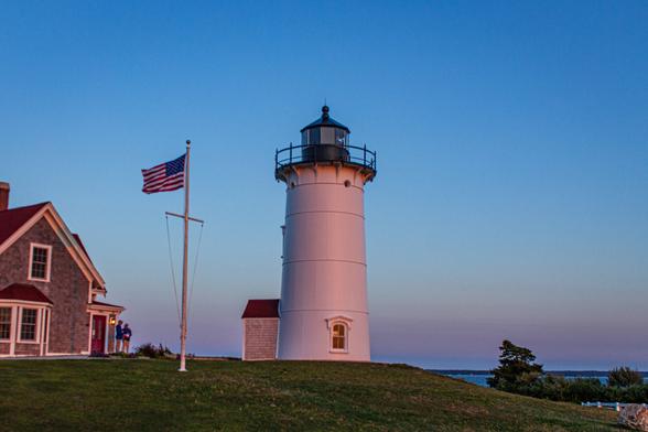 A classic white lighthouse with a black lantern room stands on a grassy hill at dusk, bathed in soft pink and blue hues from the fading sunlight. To the left, an American flag flutters on a tall flagpole beside a charming shingled house with a red roof and a warmly lit doorway. Two people in jackets stand near the house, adding a human element to the peaceful coastal scene. In the background, the ocean stretches toward the horizon, meeting the twilight sky in a gradient of deep blues and purples.