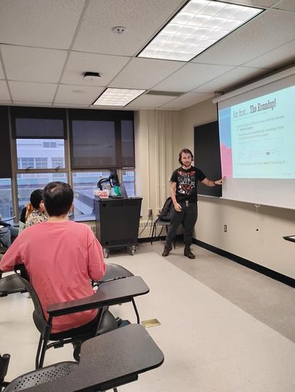 A young man with a black graphic t-shirt and sweatpants presenting a slideshow in front of a university classroom. Visible are two students in the front row. Title slide text reads: "But first, The Roundup!"