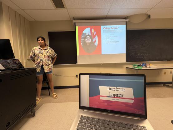 A young woman in jean shorts and a patterned button-down t-shirt presenting a slideshow in front of a university classroom. Visible is a laptop at a desk in the front row, where text reads: "Linux for the Layperson." Title text on the presenting slide reads: "Who am i?"