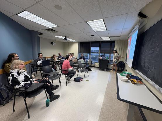 A wide shot of a university classroom, where a young woman is seen presenting to a group of students sitting at individual chairs with built-in side desks.