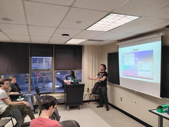 A young man in sweatpants and a graphic black t-shirt presenting a slide show in front of a university classroom. Visible is one person in the front row and two people in the row behind. Text on the presenting slide reads: "Cinnamon"