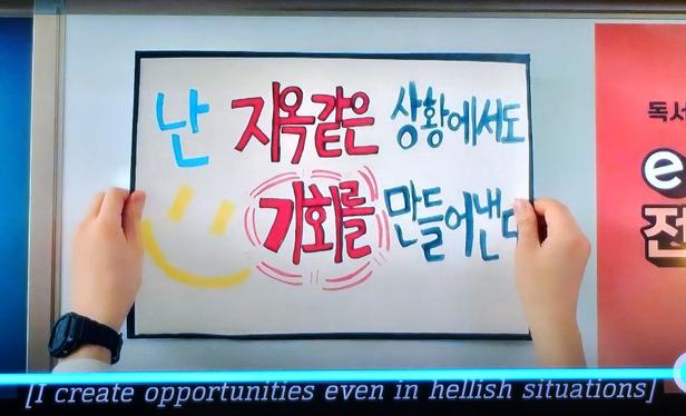 Two hands put up a poster on a wall: "난 지옥같은 상황에서도 기회를 만들어낸다" translated to "I create opportunities even in hellish situations."