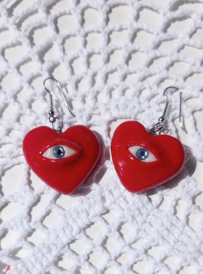 Un par de aretes con forma de corazón rojo. Cada corazón tiene un ojo en el centro, con un iris azul y una pupila negra. Los aretes están sobre una superficie blanca con un patrón tejido.