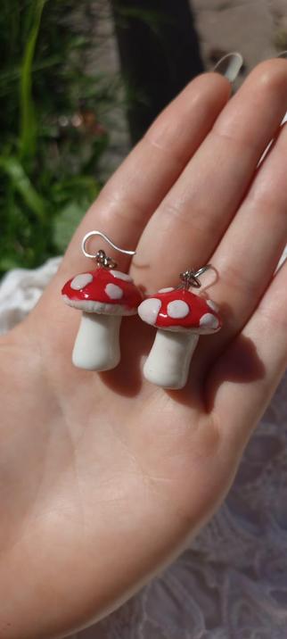Un par de aretes en forma de hongos. Los hongos tienen un sombrero rojo con puntos blancos y un tallo blanco. Los aretes están sostenidos en la palma de una mano. El fondo es borroso, pero se pueden distinguir algunas plantas verdes.
