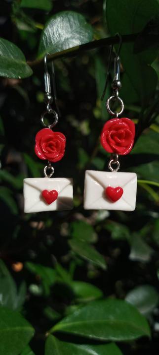 Un par de aretes colgantes con un diseño romántico. Cada arete tiene una rosa roja en la parte superior, seguida de un pequeño sobre blanco con un corazón rojo en el centro. Los aretes están colgados de un gancho de metal y están fotografiados frente a un fondo de hojas verdes.