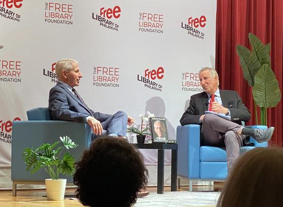 Two white-haired white men in suits sit in sky blue modern armchairs in front of a screen that reads "Free Library of Philadelphia" in a repeating pattern.  There are also a few fake plants, and a red curtain occupies the right side of the photo.  Fauci is on the left and is speaking.  The backs of a couple of spectator heads are visible at the bottom of the shot.