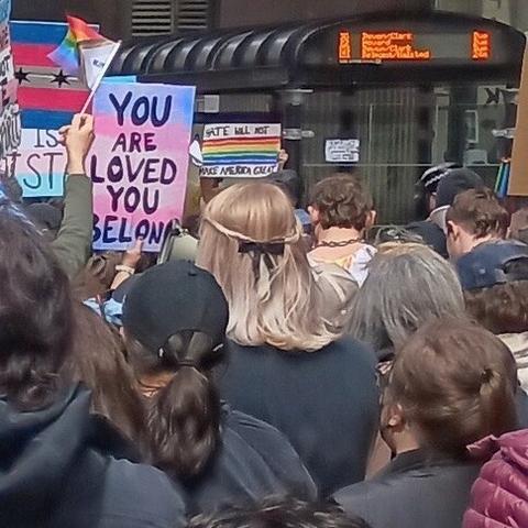 crowded chicago street, protest sign with 🏳️‍⚧️ trans colors saying “you are loved you belong,” backs of many headsand center a blonde one with a small black bow