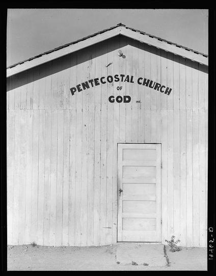 The image shows a black and white photograph of the Pentecostal Church building in Greenfield, Salinas Valley, California. It features a plain wooden structure with visible horizontal siding, a simple gabled roof without any trim or decoration, and a single closed door at its center. Above the doorway are two lines of text that read "PENTECOSTAL CHURCH OF GOD INC." The photo captures only part of the building's facade against a clear sky backdrop, suggesting an arid environment as hinted by patches of dry ground visible in front of it.