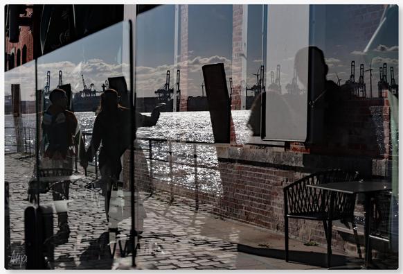 Blick auf eine Scheibe in der sich Fußgänger, Hafenkräne und funkelndes Wasser der Elbe spiegeln. Die Scheibe schützt vor Wind und dient als Absperrung für ein Cafe. Dahinter kann man Stühle und Tische erahnen und eine große Fensterfront des Gebäudes ist zu sehen. Die Farben sind gedämpft, blau-grau und rot-braun.

View of a pane in which pedestrians, harbour cranes and the sparkling waters of the Elbe are reflected. The pane protects against the wind and serves as a barrier for a café. Behind it you can see chairs and tables and a large window front of the building. The colours are muted, blue-grey and red-brown.