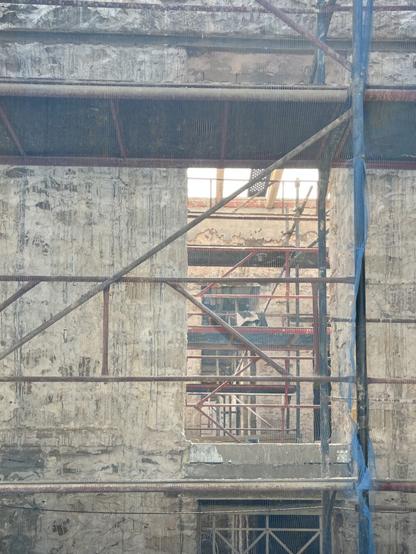 The shell of a building in Petralona, covered in scaffolding and blue cloth. The cloth is transparent from this angle, just a blue mist over the pale greyish ochre building and rust and black scaffolding.
Through an empty window, more scaffolding, more windows beyond, sunlight and the roofless sky. 
There’s a repeating pattern to the scaffolding framework seen at different distances that gives something of the air of an abstract painting, a study in parallels, diagonals, and right angles.