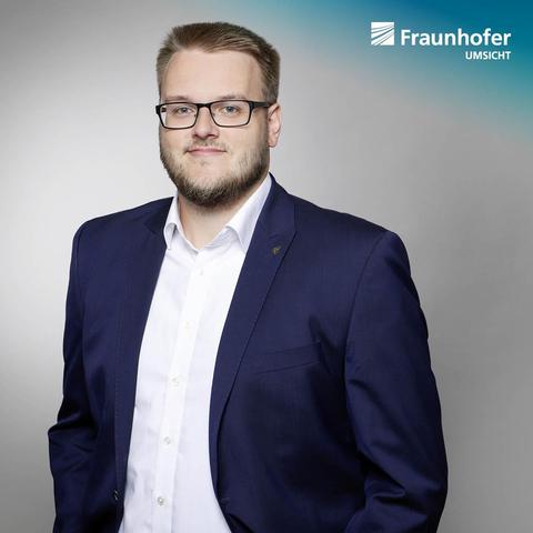UMSICHT-Wissenschaftler Kai junge Puring trägt einen dunkelblauen Anzug mit einem weißen Hemd und steht vor einem neutralen Hintergrund. Das Bild ist mit dem Logo von Fraunhofer UMSICHT in der oberen rechten Ecke versehen.