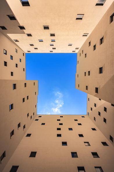 Look up view to a blue sky with a little rising white cloud from modern and geometric inner courtyard at Stralauer Platz in Berlin. The warm tones and square/solid  shapes in the houses forming the square form a nice contrast with the sky and the lightness of the clouds.