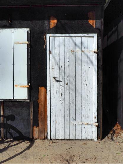 Besonnt wird eine weiße Schuppentür und ein geschlossener Fensterladen. Ins Bild fällt der Schatten eines Fahrrades.