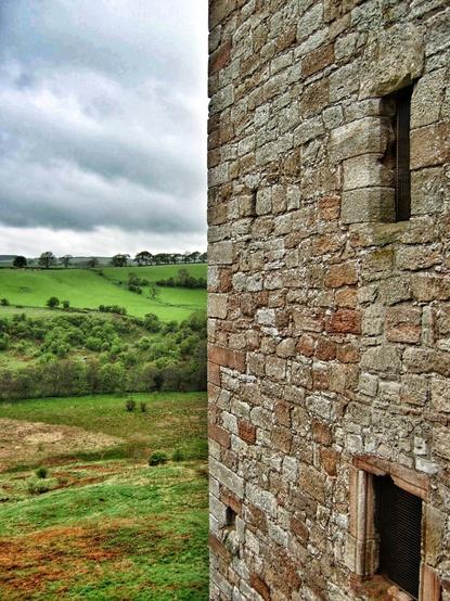 A close-up, slightly high-angle shot shows a weathered stone wall with two small, barred windows on the right side of the frame. To the left, a vast, undulating green landscape stretches out under a cloudy sky. The stone wall is constructed from irregularly shaped, light brown and grey stones, showing signs of age and weathering. The windows are small, rectangular openings with dark metal bars.
The landscape on the left is a patchwork of vibrant green fields and darker green patches of trees and bushes, suggesting rolling hills and valleys. A line of trees is visible on the horizon under a heavy, overcast sky filled with grey clouds. The overall lighting suggests a daytime scene under cloudy conditions.
