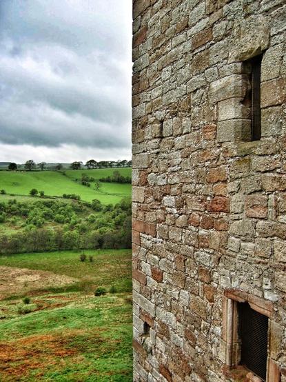 A close-up, slightly high-angle shot shows a weathered stone wall with two small, barred windows on the right side of the frame. To the left, a vast, undulating green landscape stretches out under a cloudy sky. The stone wall is constructed from irregularly shaped, light brown and grey stones, showing signs of age and weathering. The windows are small, rectangular openings with dark metal bars.
The landscape on the left is a patchwork of vibrant green fields and darker green patches of trees and bushes, suggesting rolling hills and valleys. A line of trees is visible on the horizon under a heavy, overcast sky filled with grey clouds. The overall lighting suggests a daytime scene under cloudy conditions.