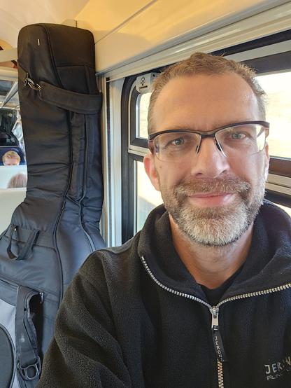 Im Vordergrund ich, lächelnd, mit schwarzer Jacke und offenem Kragen mit grauem Reißverschluss. Im Hintergrund mein Kontrabass in grauer Hülle vor einem Zugfenster