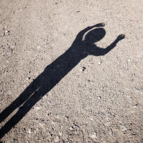 A high-angle shot captures the shadow of a person cast on a rough, light-brown surface, likely gravel or dirt. The shadow is elongated and stretches diagonally across the frame from the bottom left towards the upper right. The figure appears to be standing with their legs slightly apart and their arms raised and bent above their head, suggesting a stretching or dance-like pose. The hands are slightly spread out. The head is a solid, rounded shape. The shadow is sharply defined, indicating bright sunlight from above and slightly behind the subject. The ground is uneven with small stones and variations in texture, visible within and around the shadow.
