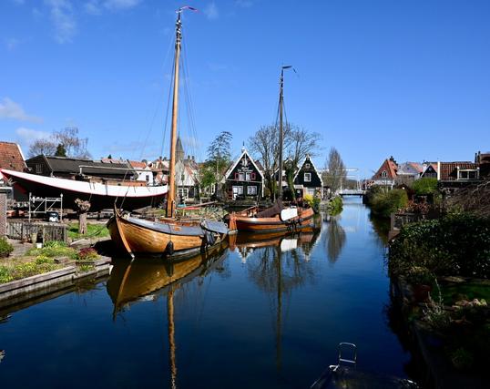 A scenic view of a canal lined with traditional wooden boats. A larger boat is on a dock for repairs, while quaint houses with varied roofs are visible along the waterway. The sky is clear with a few clouds, reflecting a serene rural atmosphere.