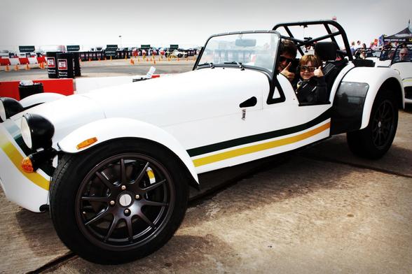 A white sports car with green and yellow stripes is parked on a concrete pavement. Two people are sitting inside the convertible, both wearing sunglasses and giving thumbs up. Orange traffic cones and multiple parked cars are visible in the background under a cloudy sky.