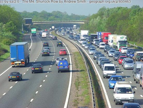 A motorway, curving sinuously through wooded terrain. On the left are three lanes of light traffic, moving easily. On the right are three lanes of bunched, crawling, dense traffic.