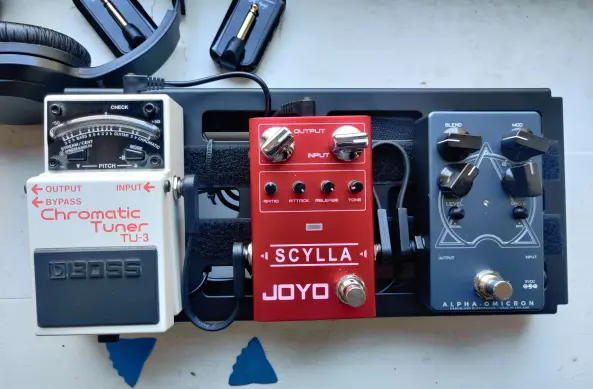 Ein kleines Pedalboard (Metall Schiene mit Durchlässen für Kabel usw, auf die man Effektgeräte für Instrumente installieren kann), auf dem man drei Metallkisten mit Schaltern und Drehknöpfen sehen kann: Ein weißes Stimmgerät, einen roten Comressor und einen stahlblauen Verzerrer. Davor liegen zwei Plectren dahinter sieht man einen Kopfhörer und zwei kleine Funkgeräte für Gitarren.