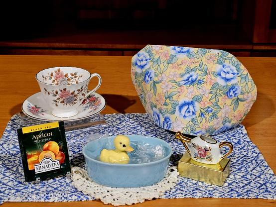 Tea tableau centered by a blue bathtub containing a small yellow duckling figurine, a porcelain watering can to refill the tub, Apricot Sunrise black tea from Ahmad, a crocheted bath mat, floral tea cozy, and flowery vintage china on a Delft blue cloth that resembles an Edwardian carpet.