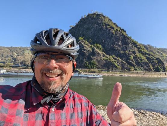 Marcus in Fahrradkleidung mit Helm und Velobrille zeigt die Daumen hoch. Er steht am Ufer des Rhein gegenüber des berühmten Loreley-Felsens. Auf dem Rhein ist noch ein Frachtschiff zu sehen.