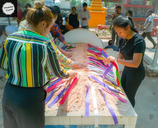 Photo shows four Khmer ladies decorating a top of a horizontal grave tomb with colourful paper strips. In the background a group of other people are preparing a show of gifts of food, drinks, valuables in honour of the ancestors. In the background other graves of other types, like the square stupa and richly decorated taller tombs. The mood is proud and grateful to celebrate the ancestors and their significance for the family and friends gathering to clean and decorate.