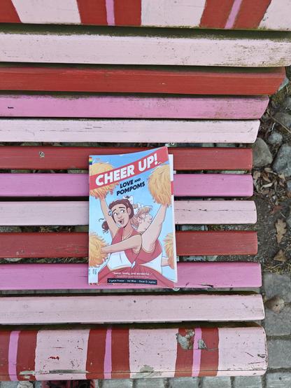 Kuvassa Cheer Up -kirja kuvattuna pinkkipunaisella puiston penkillä.