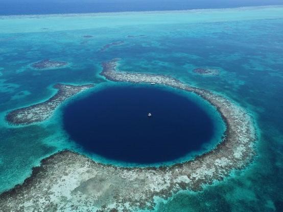 Drone footage photo taken from a height of 200 metres, looking east, showing the Great Blue Hole with the drilling platform anchored in the middle. The water is very dark blue in a round shape with turquoise colours of land and coral around it. The windward edge of the Lighthouse Reef can be seen in the background. Image thanks to E. Gischler