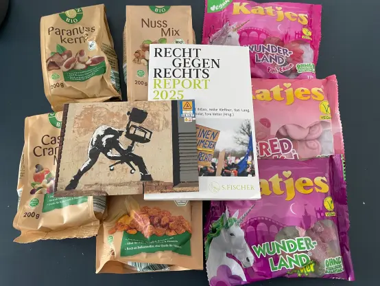 Auf einem Tisch liegen drei Packungen vegane Weingummis von Katjes, verschiedene Nussmischungen, der Recht gegen rechts Report 2025 und darauf eine Grußkarte, auf der ein Gemälde von Banksy abgebildet, dass eine Person zeigt, die einen Schreibtischstuhl so in Händen hält, als würde sie ihn zerschmettern wollen