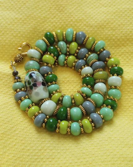 A handmade lampwork glass bead necklace  in shades of green and grey, set against a yellow fabric background.